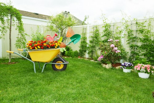 A well-maintained residential garden in Hounslow featuring a lush, green lawn bordered by a white fence, with an assortment of plants and flowers in bloom along the edges. In the foreground, a yellow garden wheelbarrow filled with brightly coloured flowers and garden tools sits on the grass, indicating recent gardening activity by Garden Maintenance Hounslow. To the right, there are flower beds with pink and purple flowering shrubs, edged with a mixture of soil and mulch. The background includes a small tree with light green foliage and several climbing plants growing along the fence, adding natural privacy and greenery to the outdoor space. The garden appears to be under mild weather conditions with natural daylight illuminating the scene, suggesting a pleasant and inviting environment suitable for outdoor gardening and landscaping services.