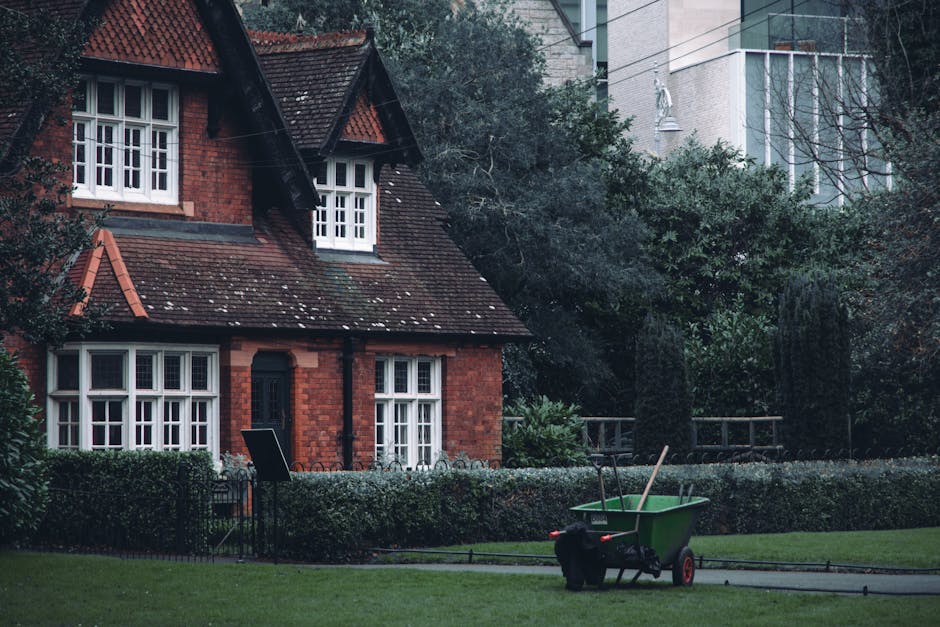 A red brick house with white-framed windows and a steeply pitched, tiled roof is surrounded by a neatly maintained garden. The garden features a lush green lawn bordered by a low hedge, with mature tr