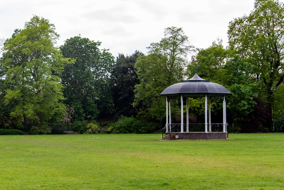 A peaceful park scene featuring a circular bandstand with a grey, domed roof supported by white columns, situated on a grassy lawn. The surrounding landscape includes neatly trimmed green grass, matur